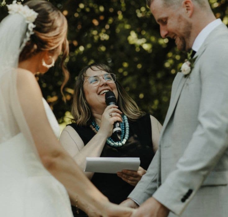 Officiant Katie Brennan standing between flower-filled milk churns in a golden meadow