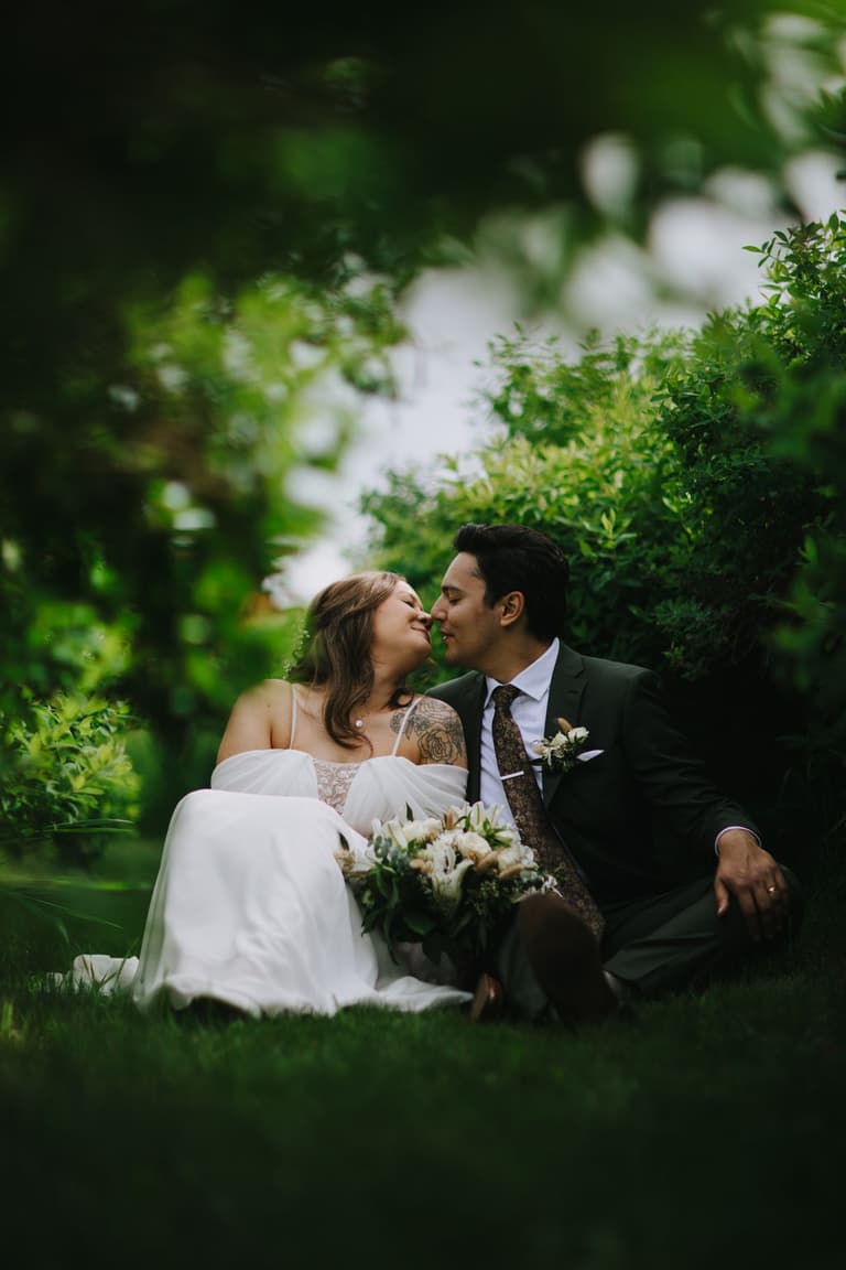 Bride and groom touching noses in garden, bride holding white bouquet, soft green foliage behind