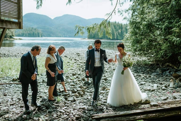 Newlyweds walking a rocky shoreline with mountains and a wedding party behind