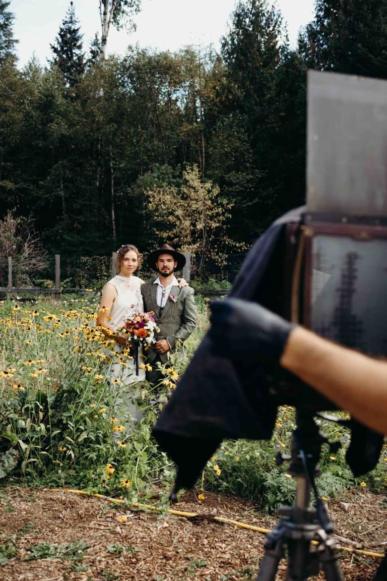 Groom in gray suit and black hat stands among wildflowers and tall trees during outdoor forest wedding ceremony