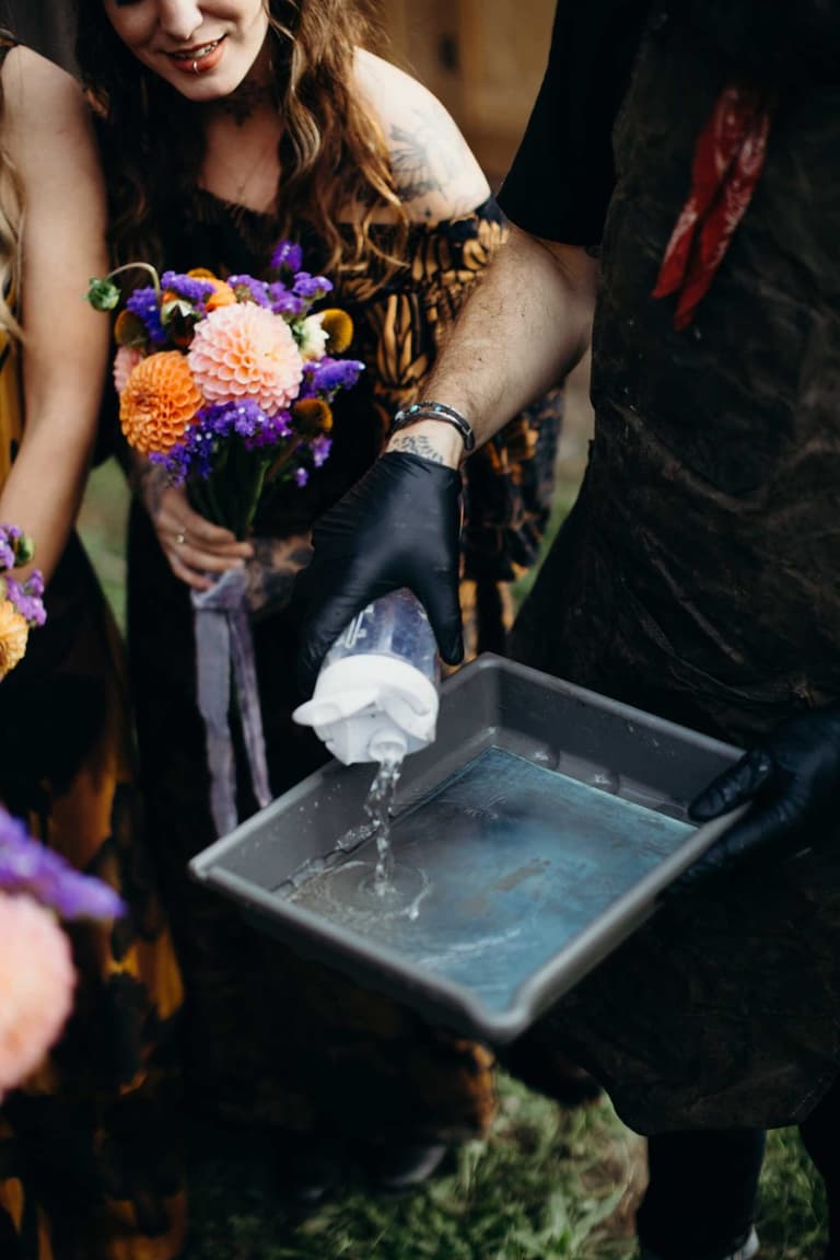 Groom in black suit and white glove pours water over bride's hand above a stone bowl, purple and yellow flowers nearby