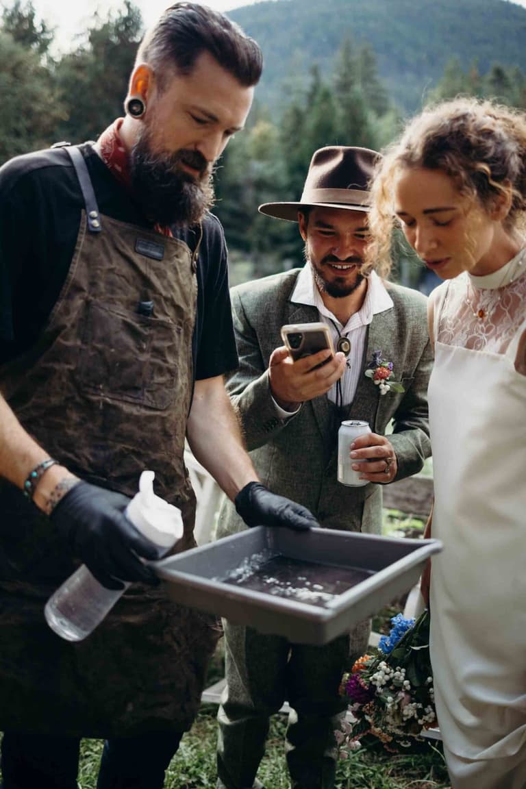 Two bearded men examine a smartphone together outdoors with forested mountains behind them