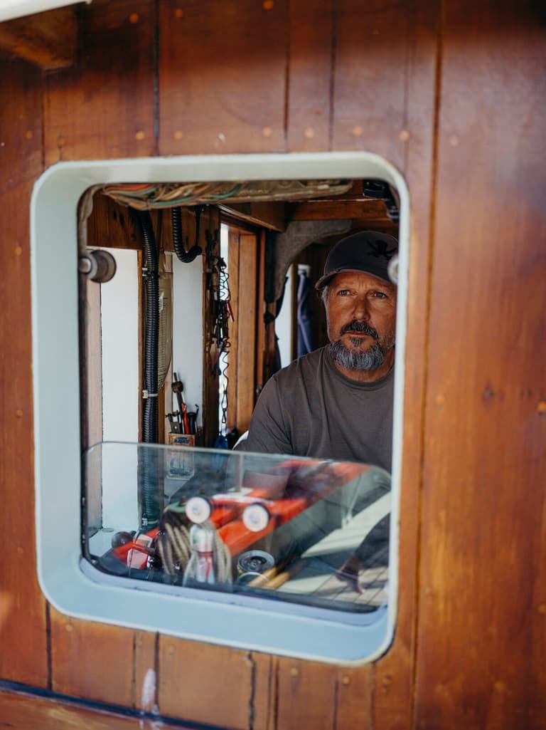 Bearded man inside wooden sailboat cabin, looking toward camera with rigging and maritime equipment visible