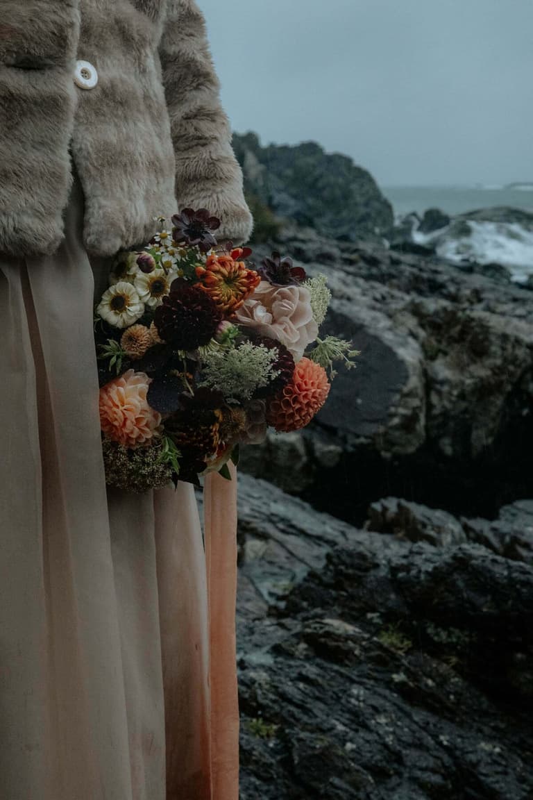Autumn dahlia bouquet held against a fur coat on rocky shoreline