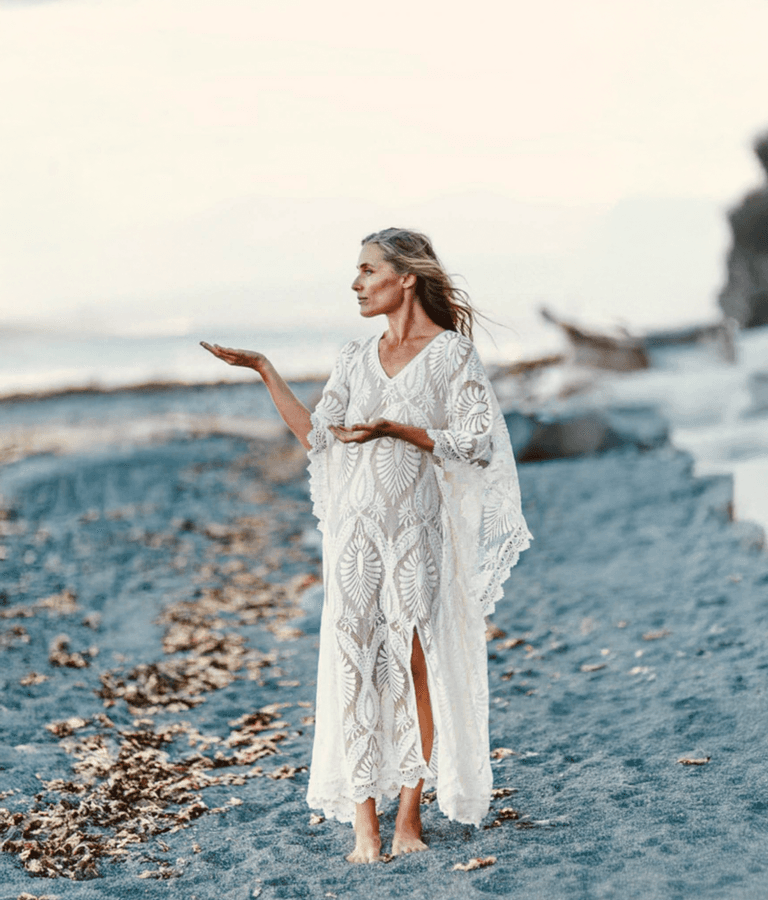 Alli Mary in a white lace kaftan on a driftwood-strewn beach
