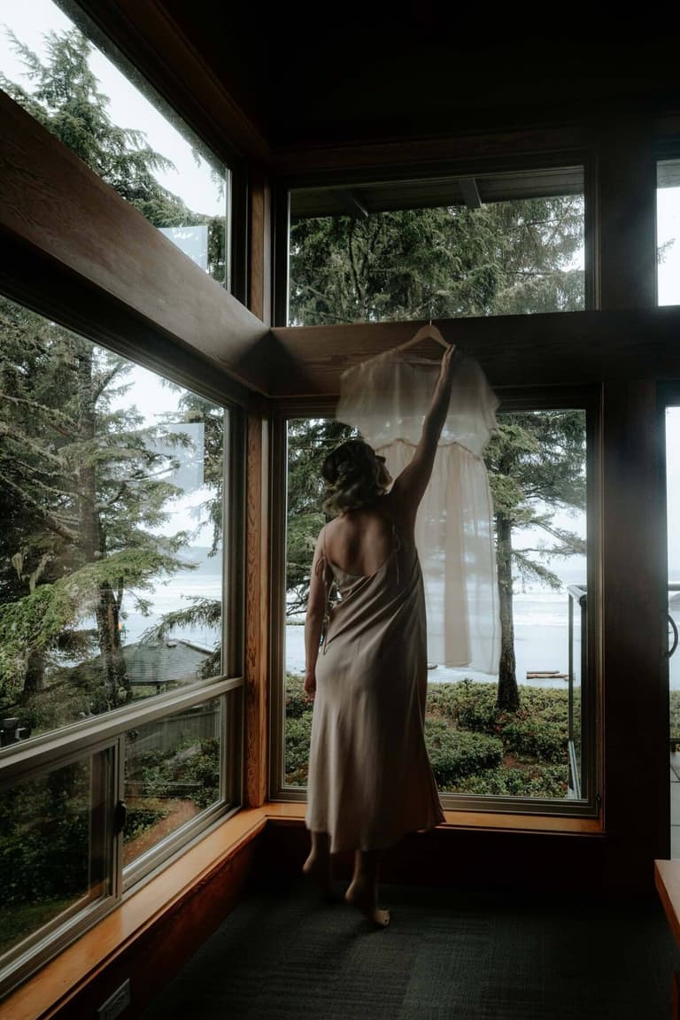 Bride reaching for her hanging dress by an ocean-view window
