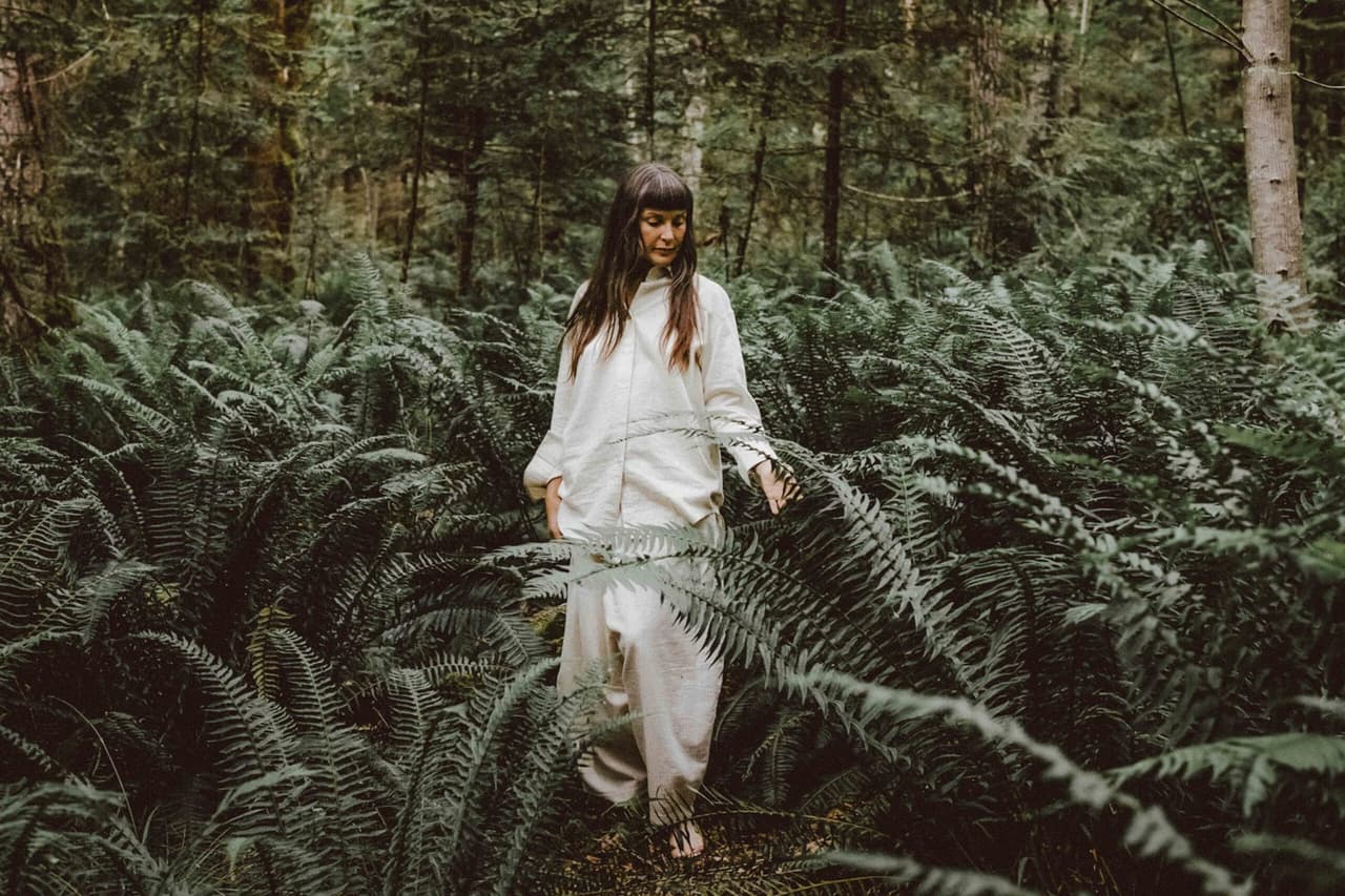 Woman in cream linen shirt and pants touches fern fronds in a mossy forest, looking down thoughtfully among green trees.