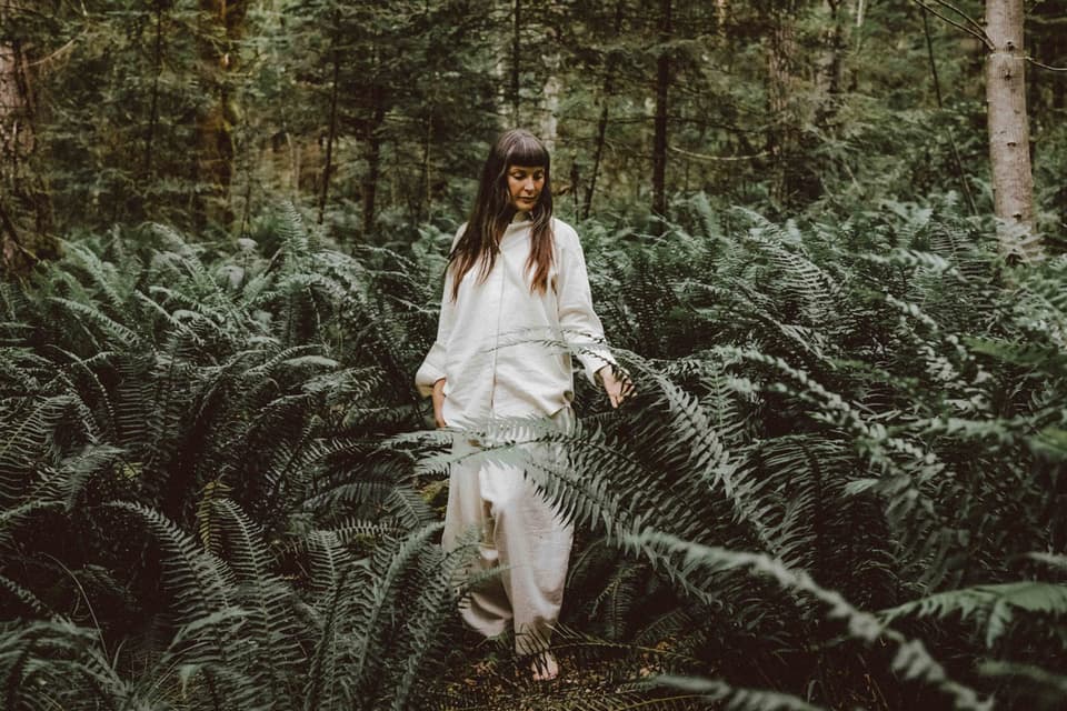 Woman in cream linen shirt and pants touches fern fronds in a mossy forest, looking down thoughtfully among green trees.