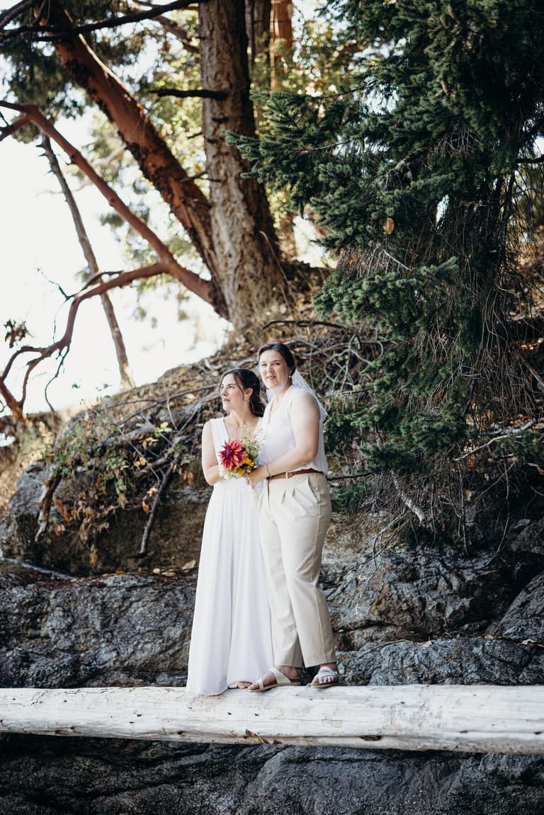 Two brides standing on a driftwood log beneath arbutus trees