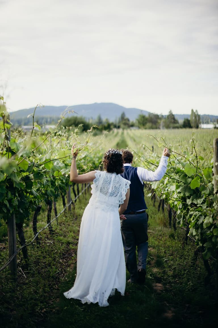 Couple raising arms triumphantly between vineyard rows, mountains beyond