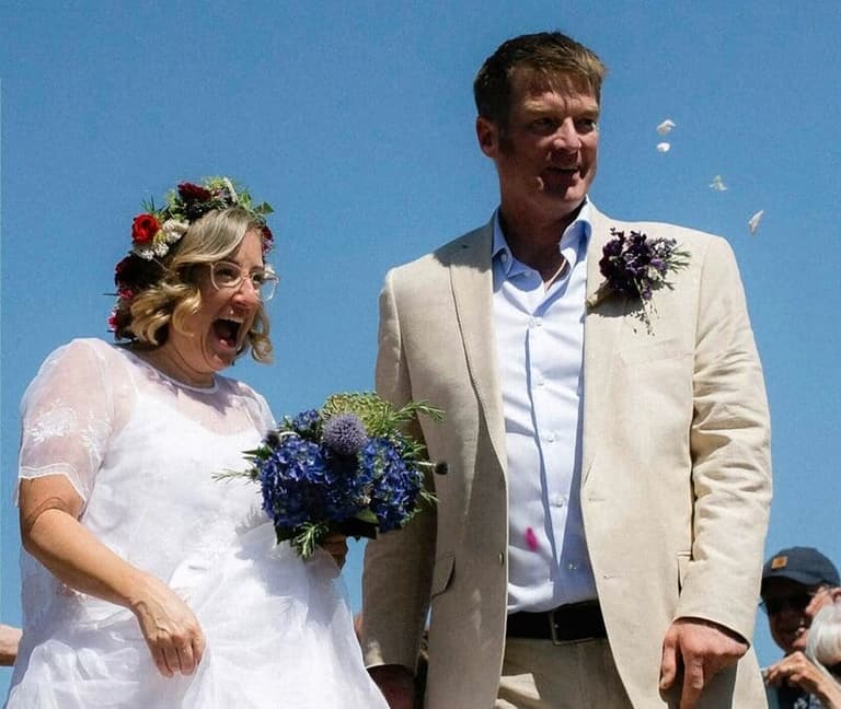 Bride in white dress with blue bouquet and groom in cream blazer smile in bright sunlight against clear blue sky
