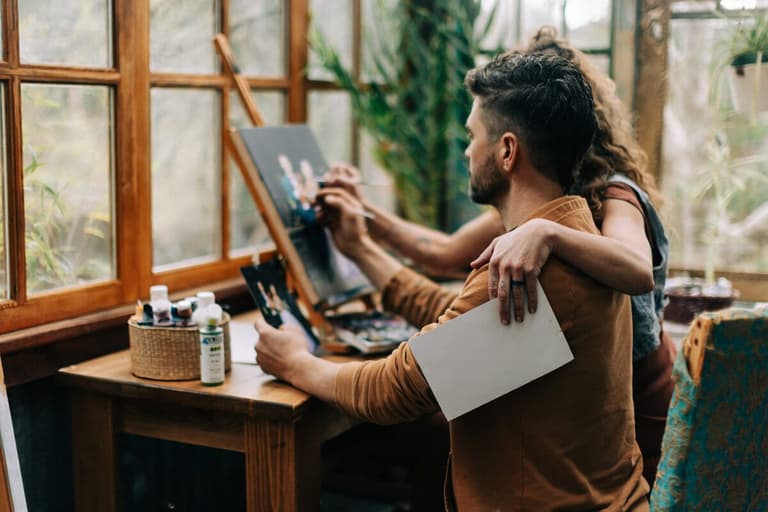 Painter at the easel with his partner's arms around his shoulders