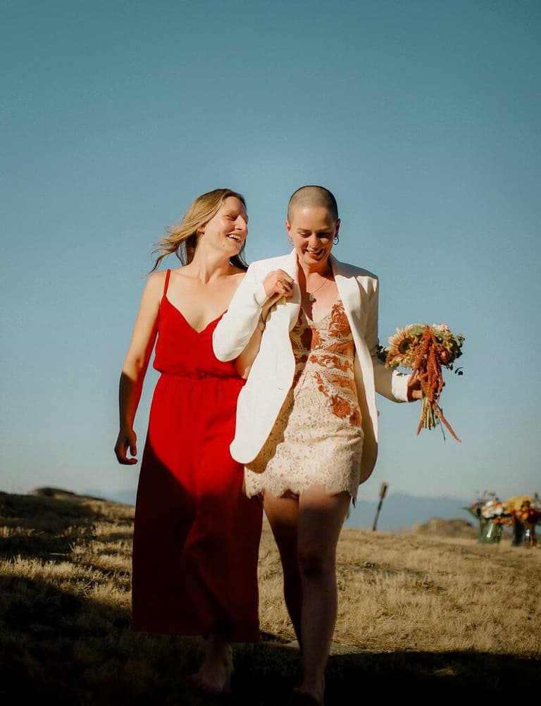 Two women walk arm-in-arm across grassland under clear sky, one holding a wildflower bouquet