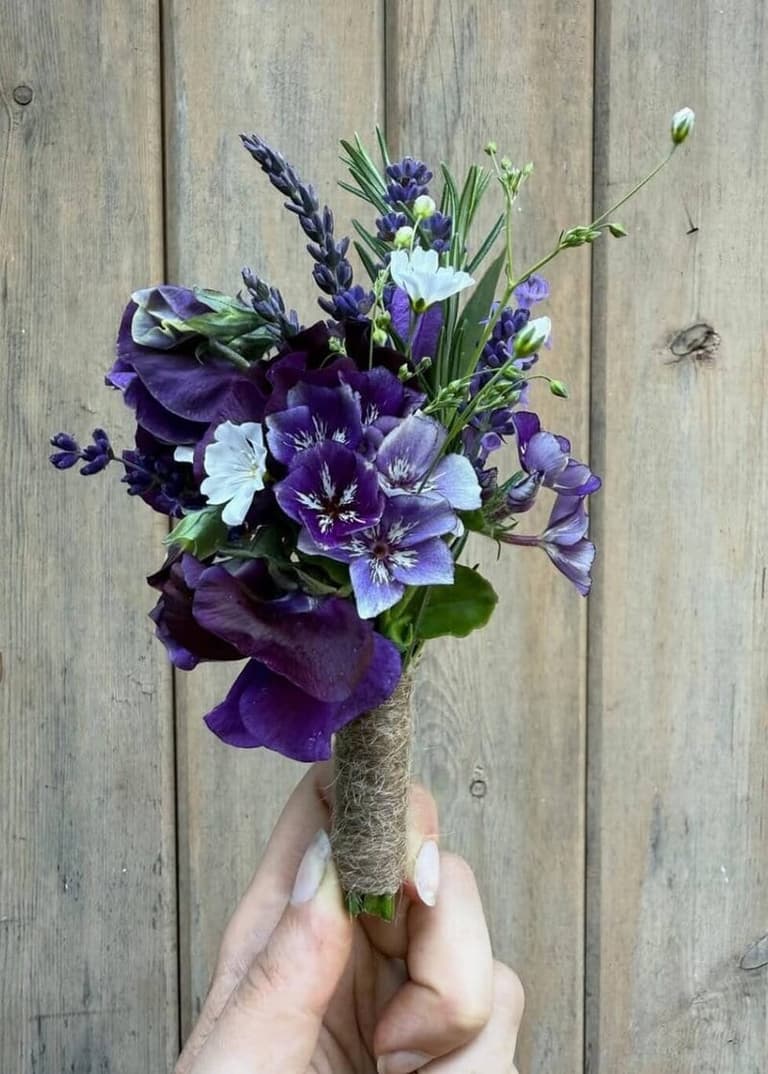 Hand holding a small bridal bouquet of purple and white flowers with twine-wrapped stem against weathered wood background