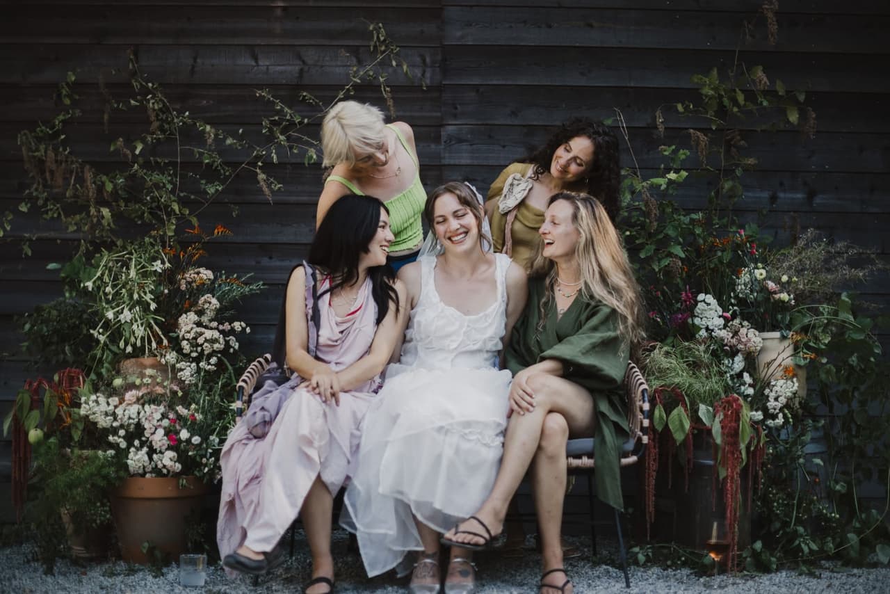 Five women laughing together against a dark wooden backdrop, bride in white dress centered among friends in pastel tones