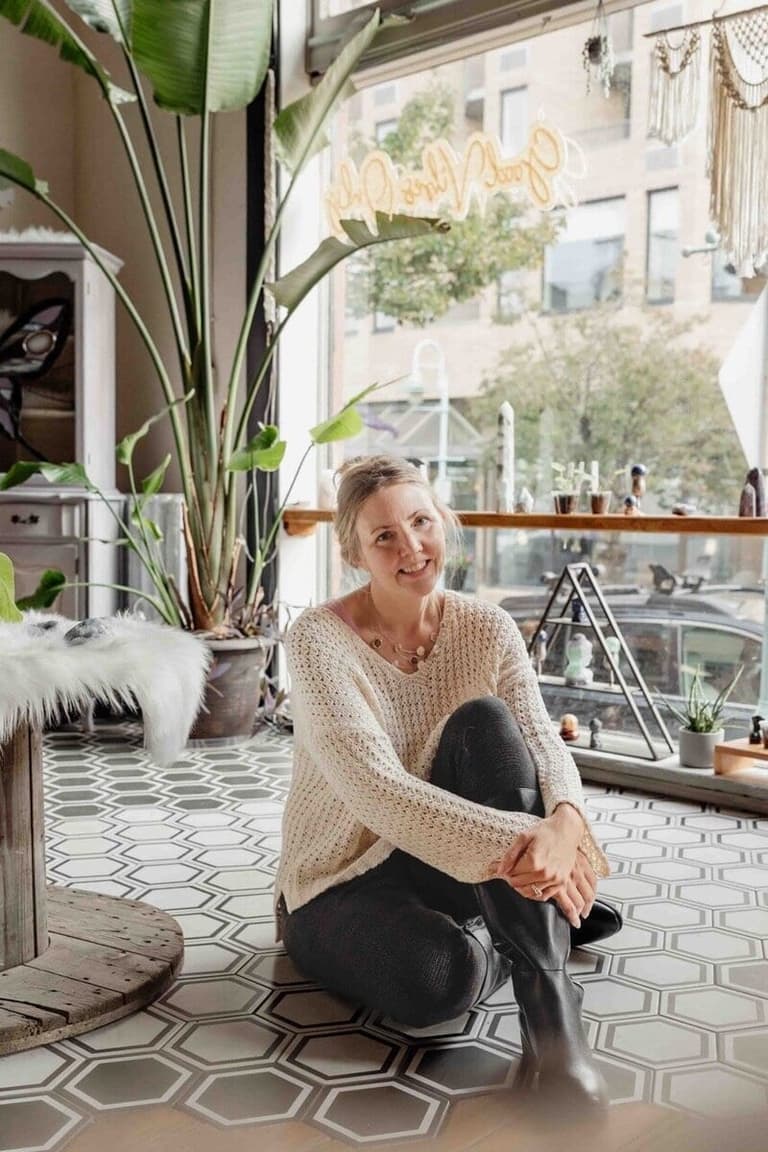 Woman sits cross-legged on geometric floor near potted plants, smiling at camera in bright indoor space
