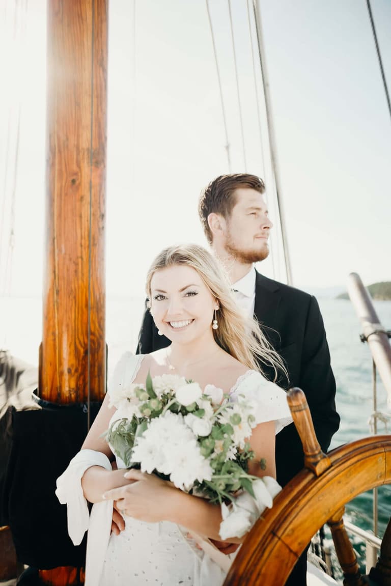 Bride holding white floral bouquet smiles while groom stands behind her aboard the sailboat wedding venue