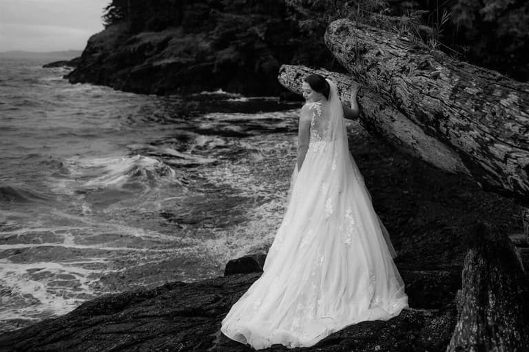 Bride in a full tulle gown on storm-washed rocks beneath driftwood