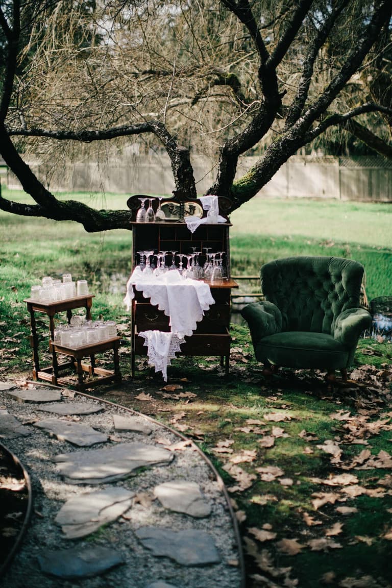 Green velvet armchair and antique glassware bar beneath a gnarled tree