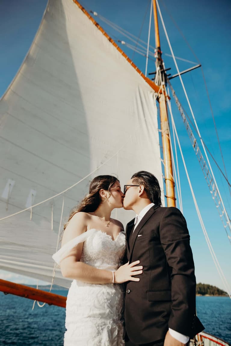 Bride and groom kiss beneath billowing white sails and clear blue sky aboard a historic tallship