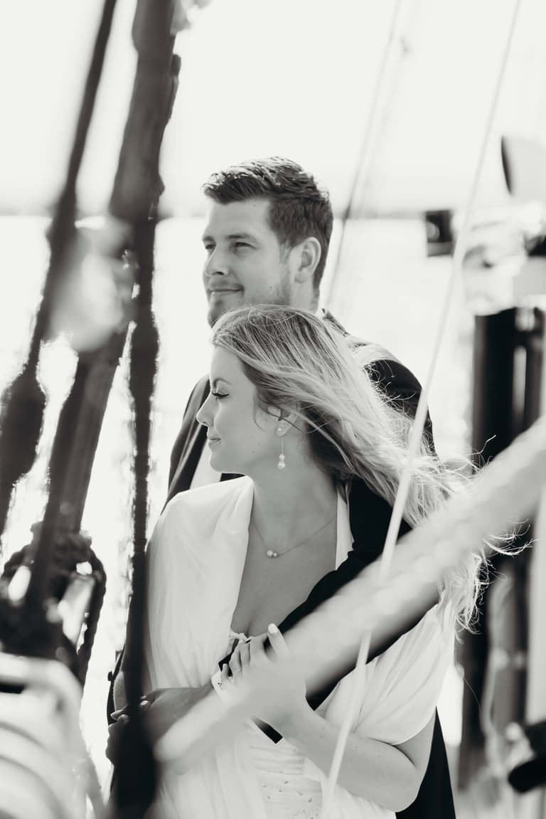 Couple in formal white attire stand close together on deck, gazing toward the horizon in black and white portrait
