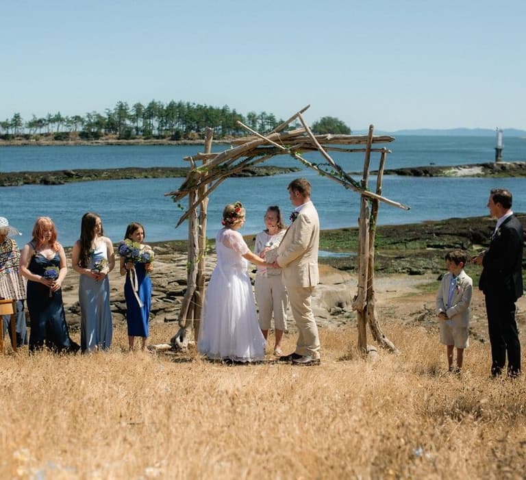 Bride and groom exchange vows beneath driftwood arbor on sandy beach with blue ocean and forested shoreline beyond