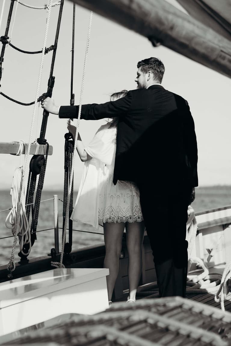 Groom adjusts white canvas sail rigging while bride stands beside him on the sailboat wedding venue deck