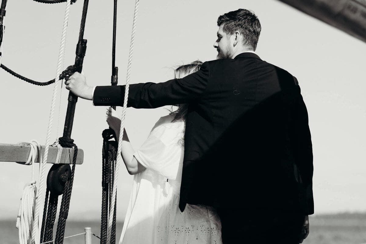 Groom reaches toward a white rope on the sailboat as bride stands beside him in profile, black and white photograph