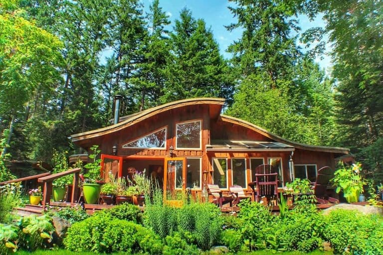Warm wood cabin with large windows nestled among tall evergreens, surrounded by lush gardens and potted plants on a sunny day