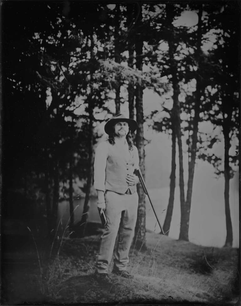 Tintype of a hatted man holding a rifle among lakeside trees