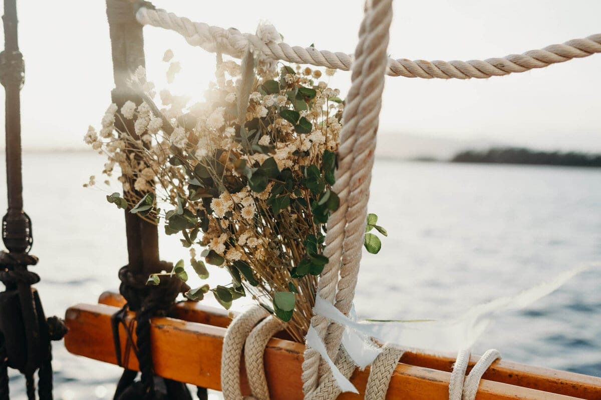 Dried flowers and green foliage tied to weathered rope rigging on a wooden sailboat, backlit by soft water and sky