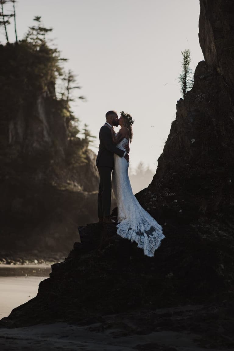 Backlit couple kissing on beach rocks, photographed by Folklore Wedding Company