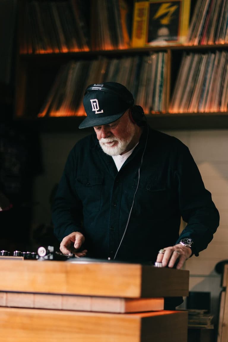 DJ in cap and headphones cueing vinyl beneath shelves of records