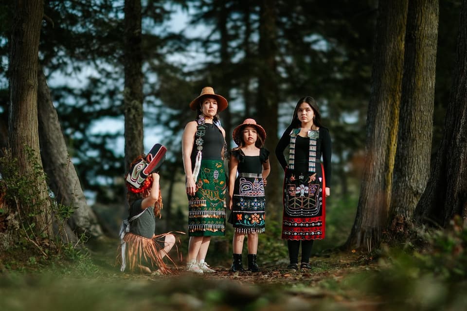 Three generations in Kwakwaka'wakw regalia standing among evergreens in dappled forest light