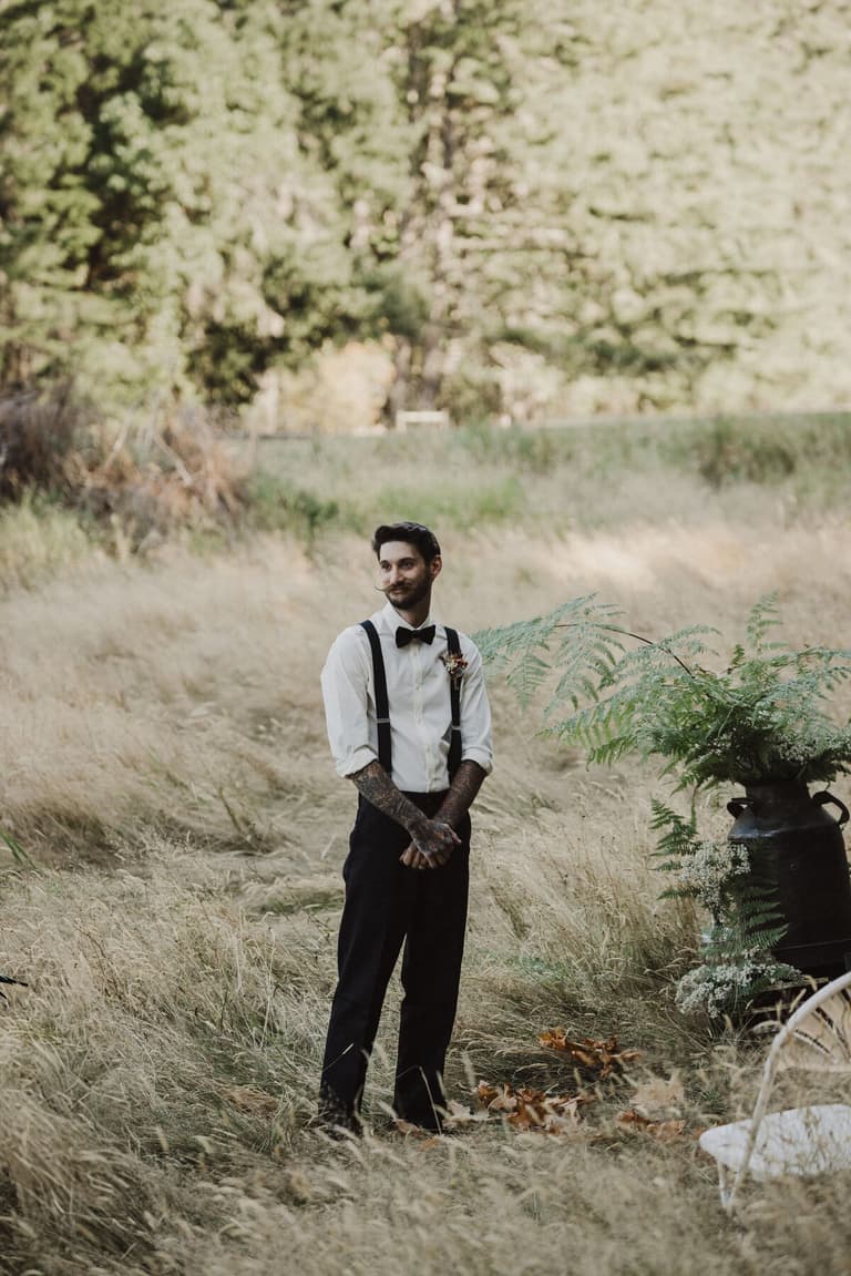 Groom in bow tie and suspenders standing in tall dry grass