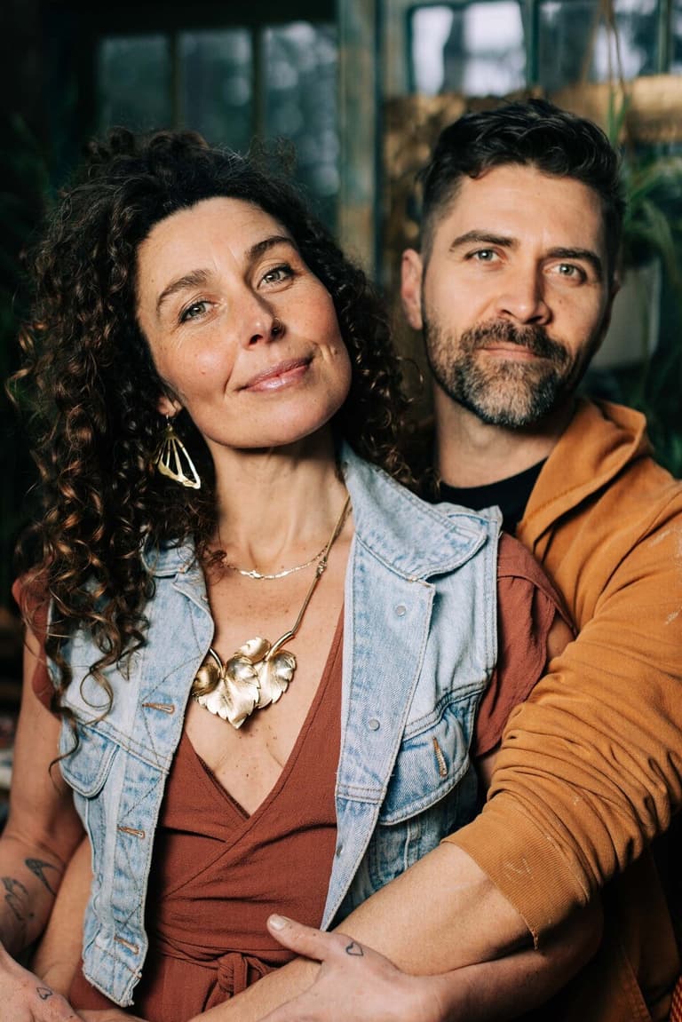 Curly-haired painter and bearded partner posing together among studio plants