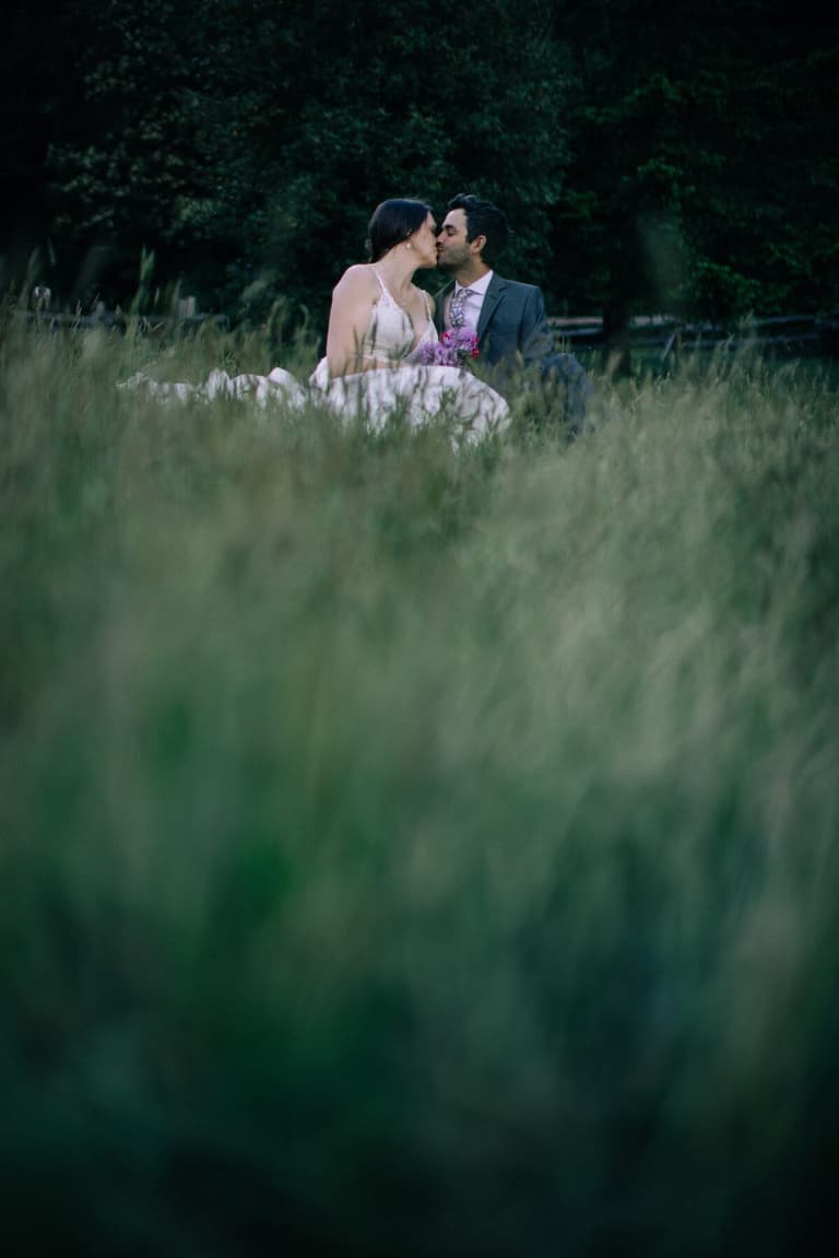 Newlyweds kissing while seated in deep meadow grass