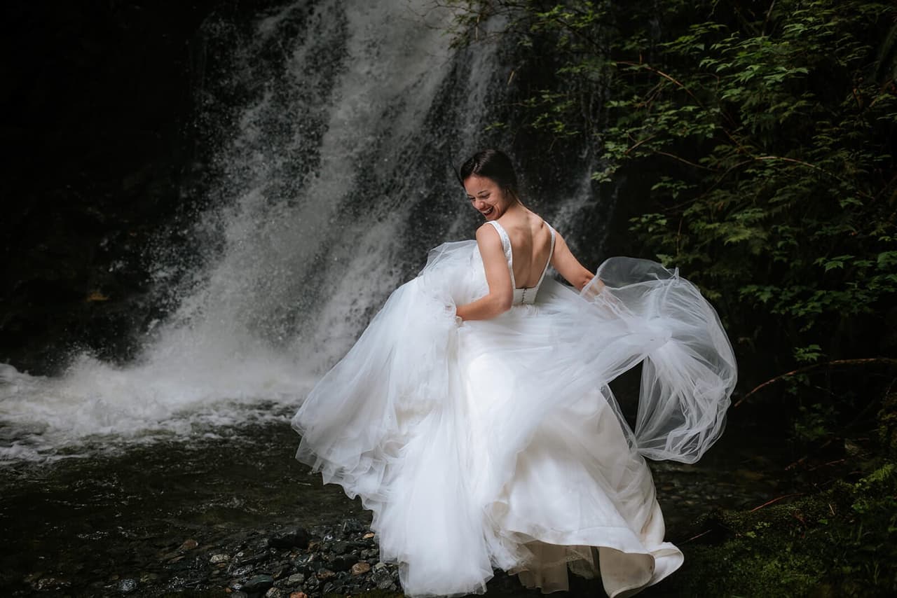 Bride in flowing white gown twirls toward weathered stone wall draped with moss at Nimmo Bay, smiling over her shoulder