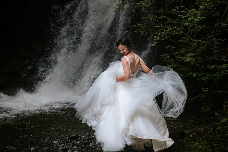 Bride in flowing white gown twirls toward weathered stone wall draped with moss at Nimmo Bay, smiling over her shoulder