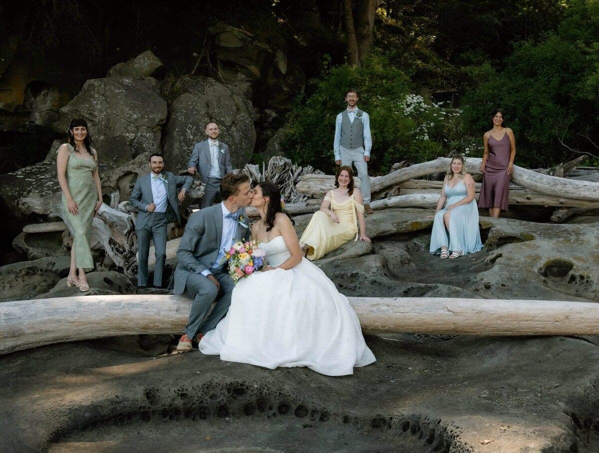 Bride and groom kiss on driftwood logs at night with wedding party standing behind them in formal attire