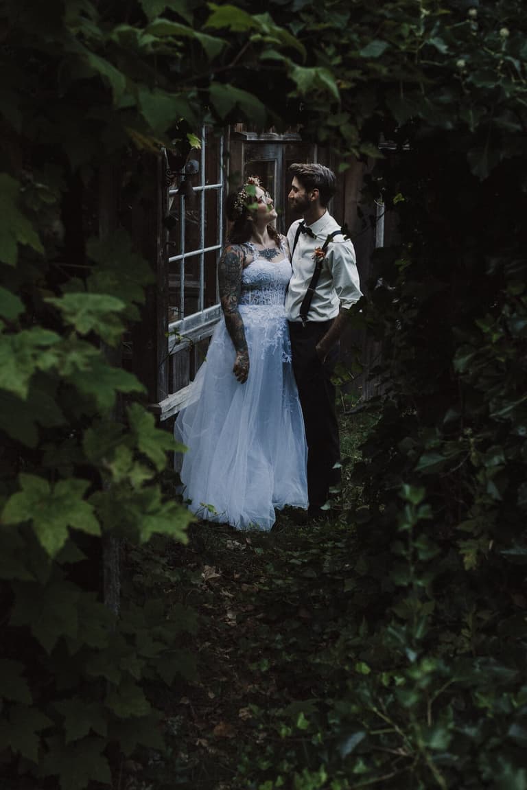 Tattooed bride and groom beside weathered greenhouse windows in deep foliage