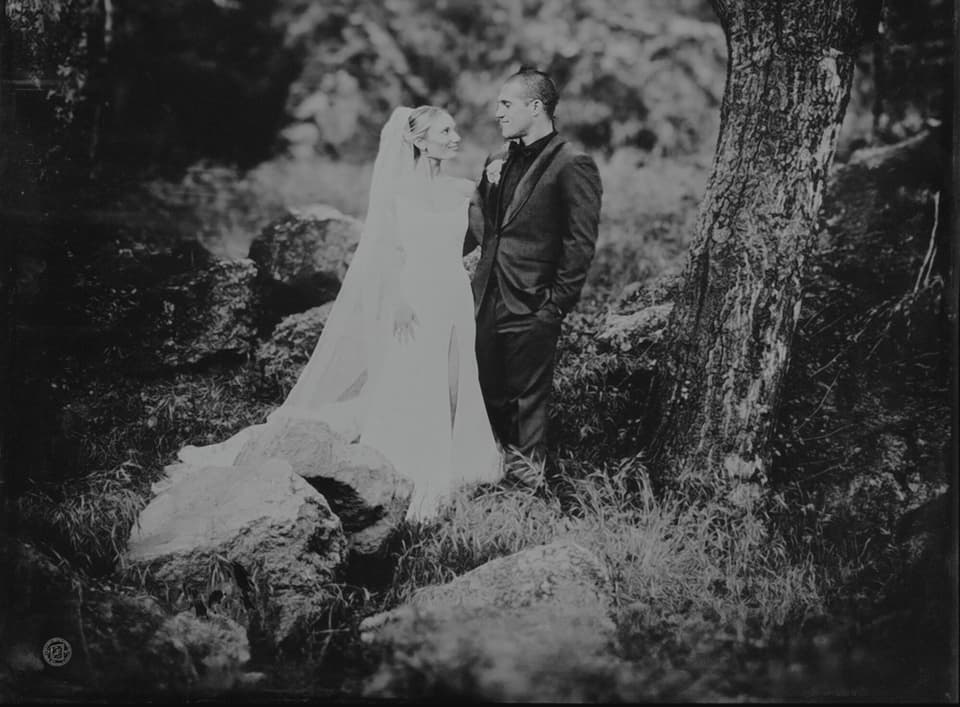 Bride in veil and groom in suit face each other outdoors, rendered as a tintype portrait with soft focus foliage behind them