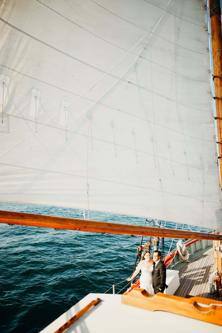 Bride seated on deck of historic wooden sailboat with full canvas sails and turquoise water behind