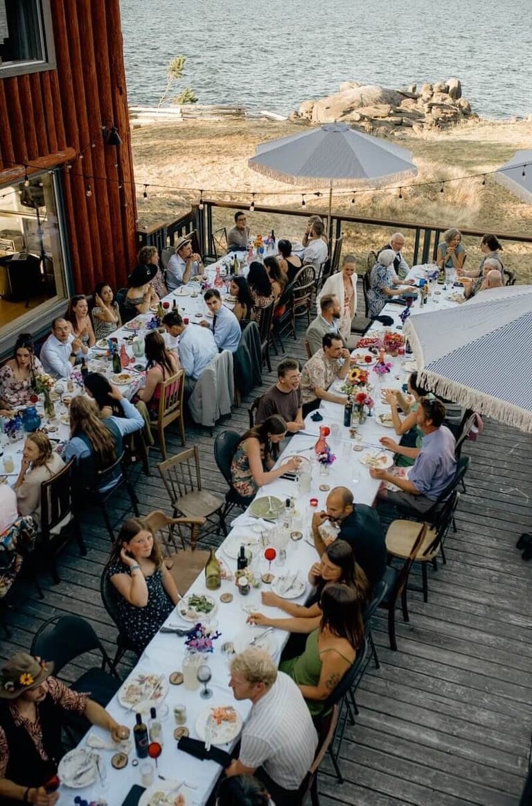Aerial view of wedding guests seated at long tables on a waterfront deck with beach and shoreline beyond