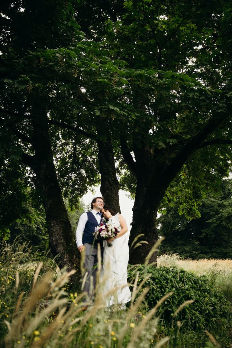 Newlyweds nestled beneath a towering tree in tall grass