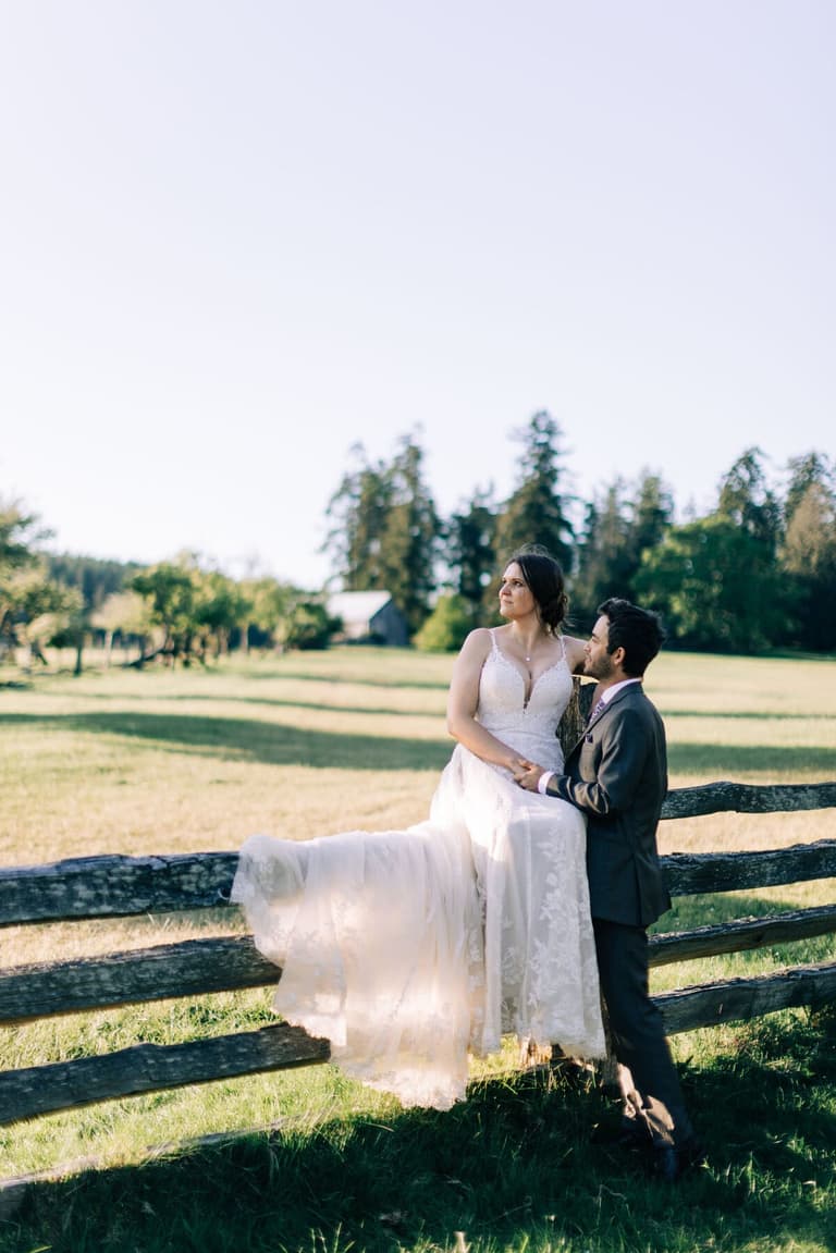 Bride perched on a split-rail fence beside her groom