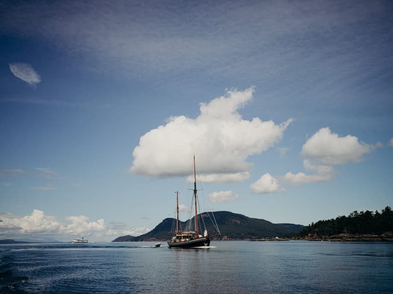 Wooden sailing vessel anchored in calm waters with forested mountain backdrop and white cumulus clouds overhead