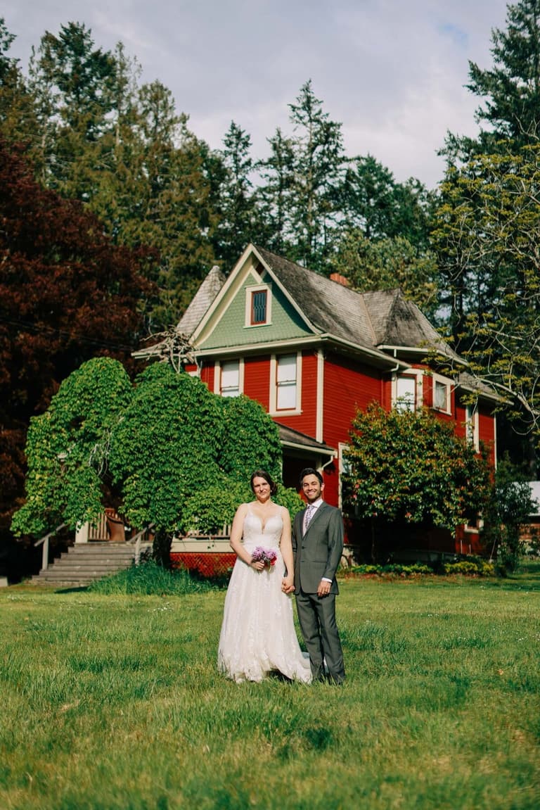 Bride and groom hold hands on the lawn in front of a red heritage farmhouse at Ruckle Farm