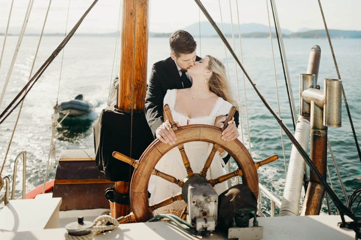 Bride and groom kiss at the wooden helm of a historic sailboat, water and distant shoreline visible behind them