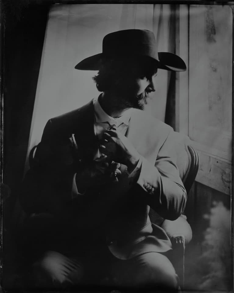 Moustached man in a cowboy hat lit dramatically beside a window